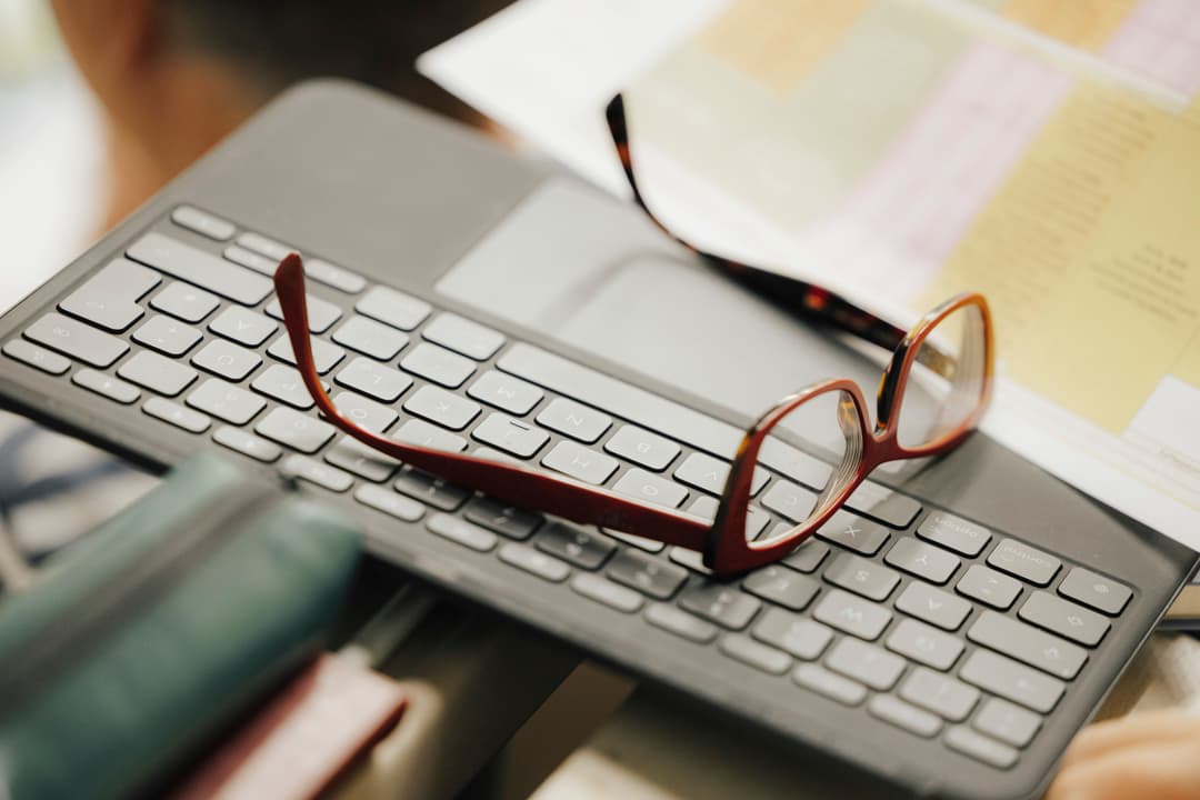 Eine Brille mit braunem Rahmen liegt auf einer modernen Tastatur mit flachen, rechteckigen Tasten. Im Hintergrund sind unscharfe Dokumente und ein weiteres Brillengestell zu erkennen. Die Szene wirkt wie ein Arbeitsplatz.